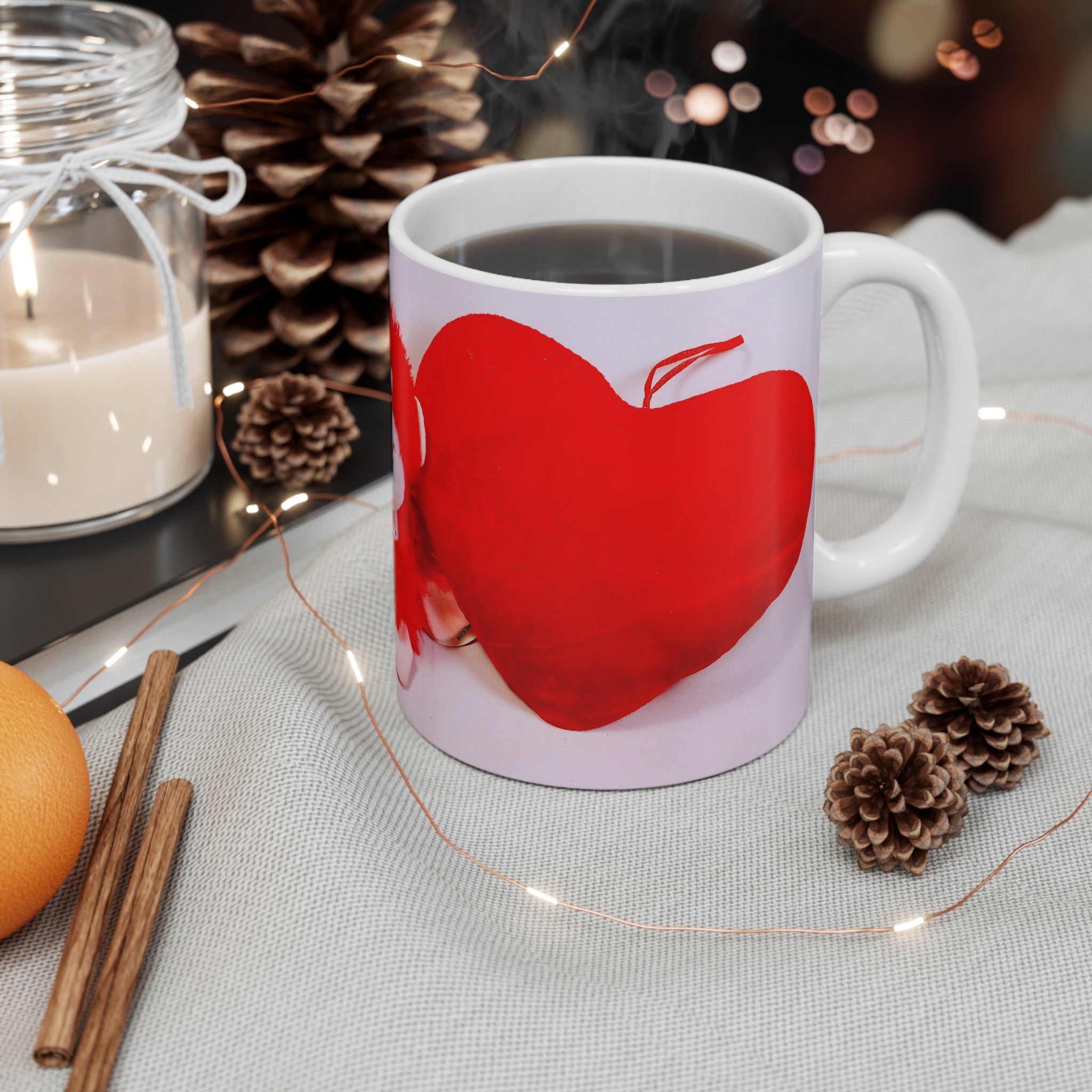 White mug with red apple design on a table with candles, oranges, and pine cones.